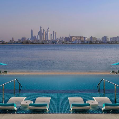 The Retreat Palm Dubai Mgallery By Sofitel Esterno foto The image shows a serene waterfront view featuring a luxurious pool area. In the foreground, there are several sun loungers and umbrellas, suggesting a relaxation space. The pool has a striking blue color, and its edge appears to blend into the body 