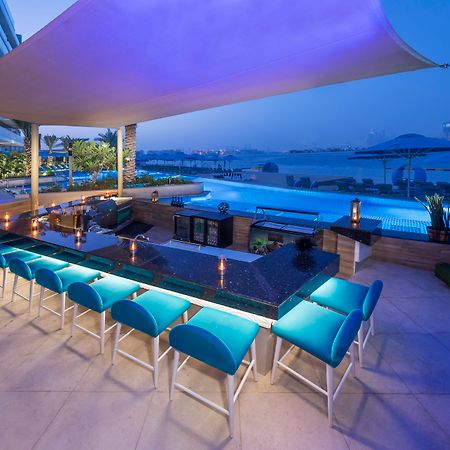 The Retreat Palm Dubai Mgallery By Sofitel Esterno foto The photo depicts an elegant outdoor bar area beside a swimming pool. It features a long, dark countertop with several bar stools that have vibrant blue cushions. The setting is illuminated with soft lighting, creating a relaxed and inviting atmosphe