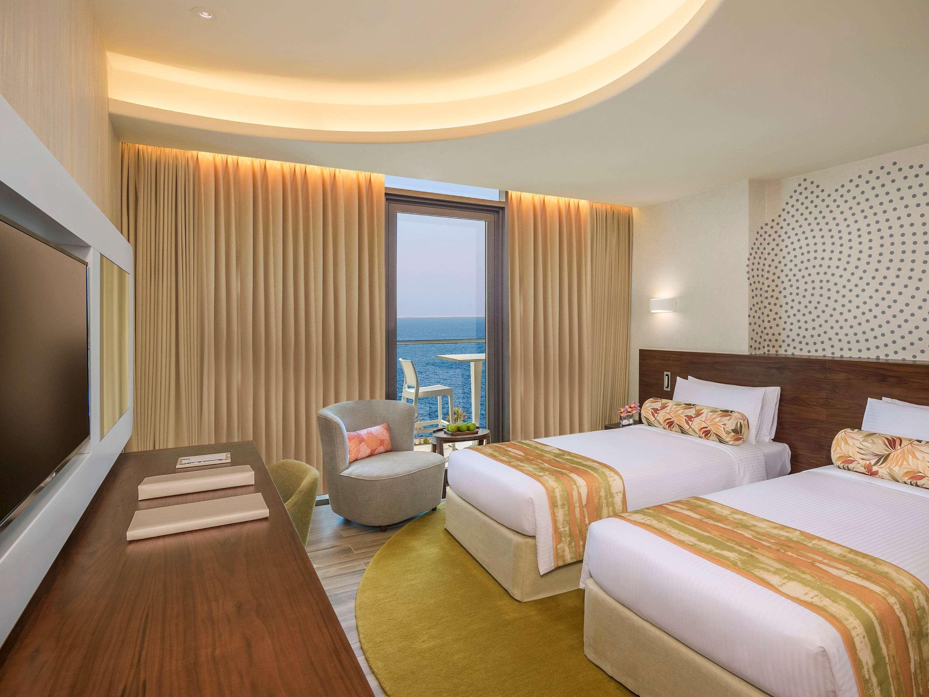 The Retreat Palm Dubai Mgallery By Sofitel Esterno foto The photo shows a modern hotel room with two twin beds dressed in white and yellow-striped blankets. There is a comfortable armchair in the corner next to a large window that offers a view of the ocean. The walls are adorned with light-colored curtai
