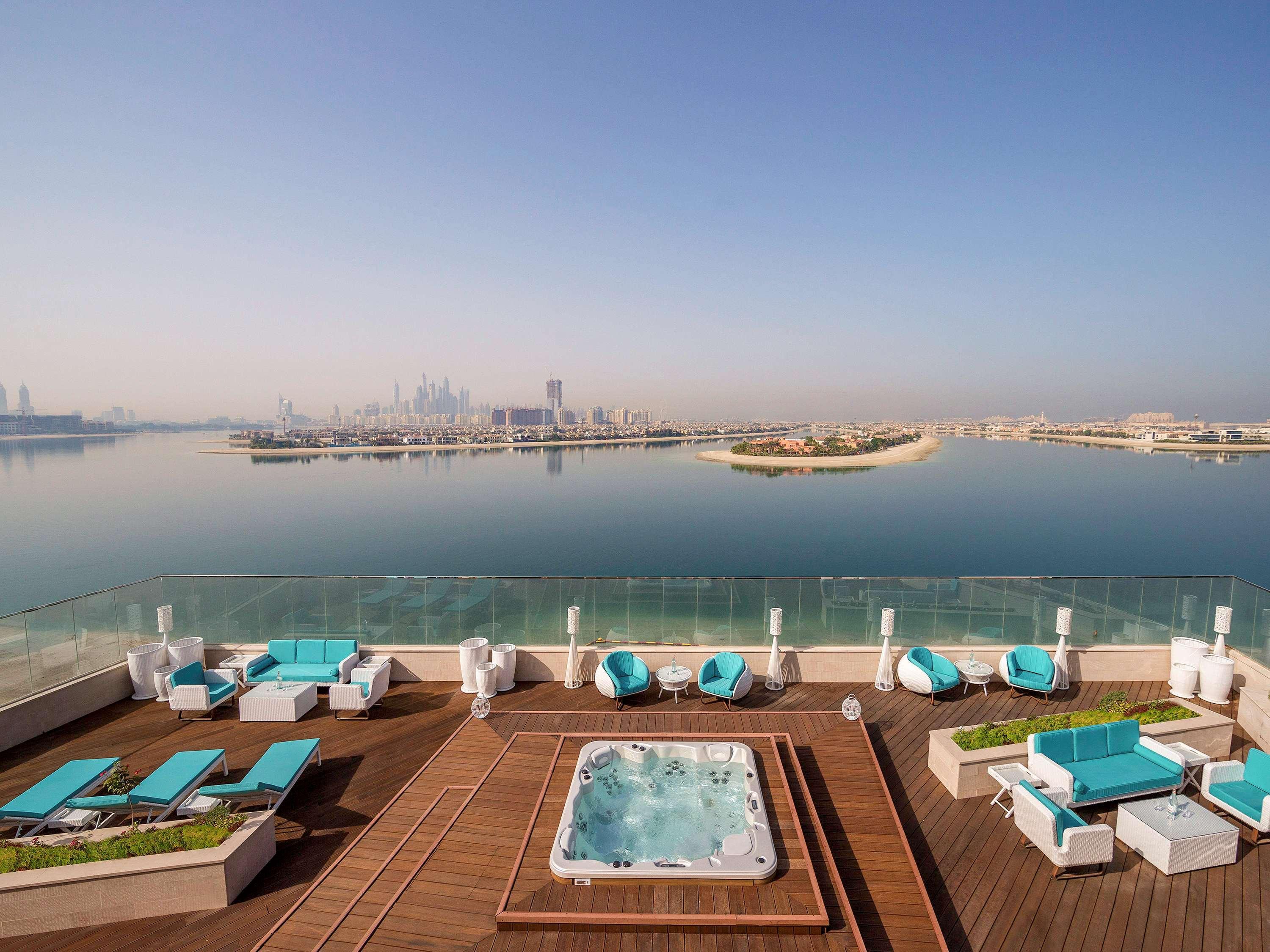 The Retreat Palm Dubai Mgallery By Sofitel Esterno foto The photo shows a rooftop area with a stunning view of a calm body of water and a city skyline in the background. There are lounge chairs and seating areas, featuring bright turquoise cushions. In the center, there is a hot tub surrounded by wooden d