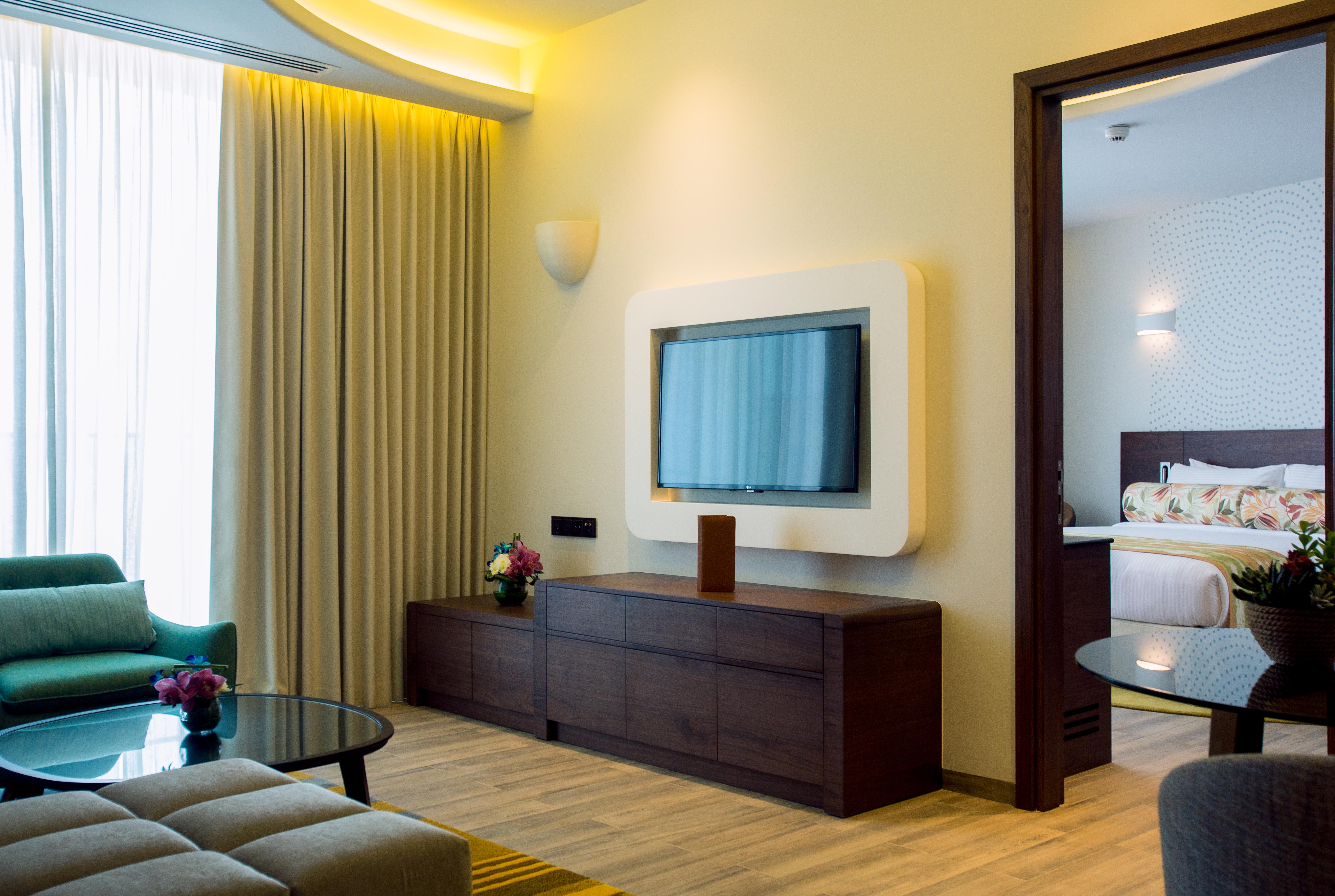 The Retreat Palm Dubai Mgallery By Sofitel Esterno foto The photo shows a modern living room area. It features a flat-screen television mounted on the wall within a light-colored frame. Below the TV, there is a sleek wooden media console. To one side, a cozy seating arrangement includes a green couch and 