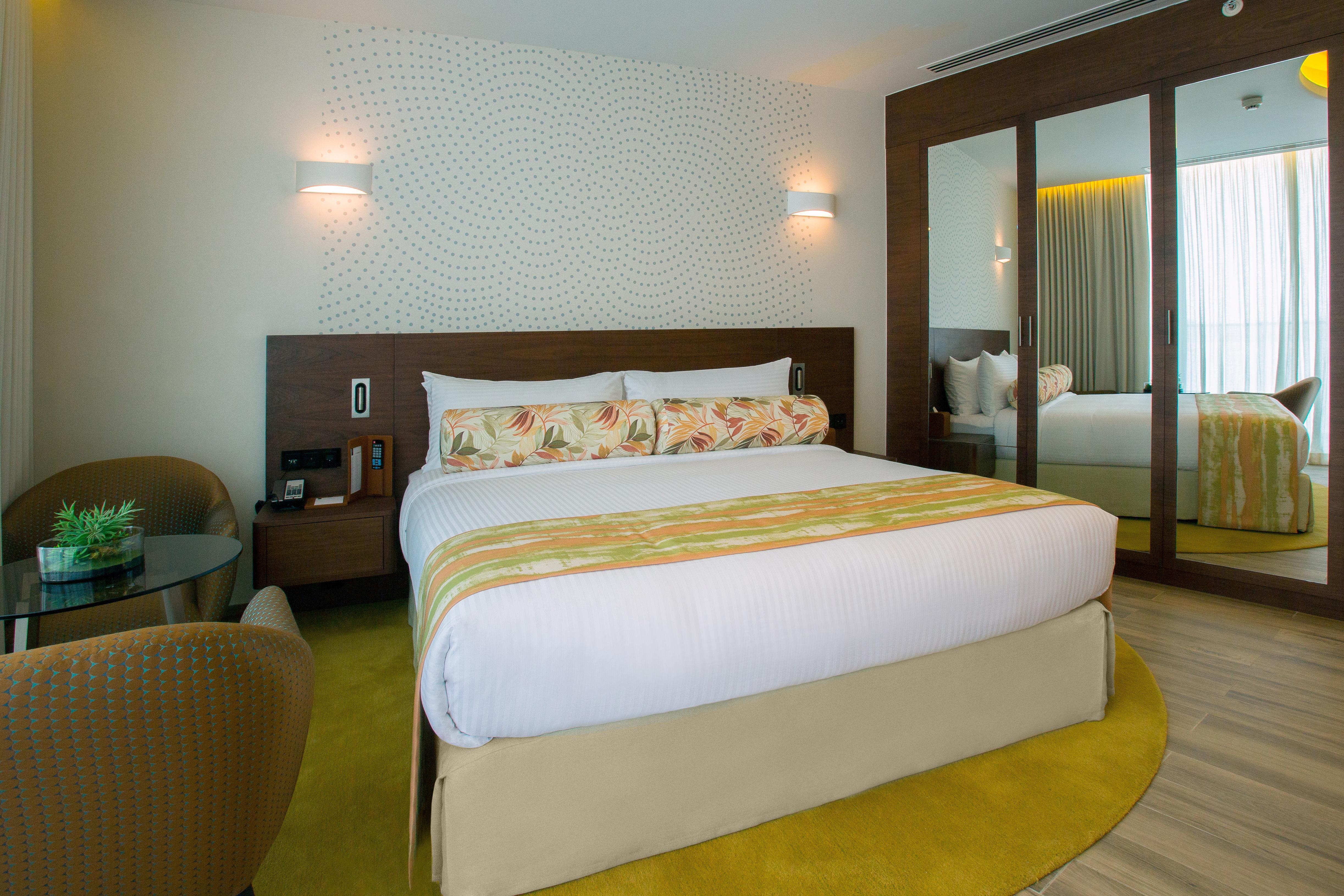 The Retreat Palm Dubai Mgallery By Sofitel Esterno foto The photo shows a modern hotel room featuring a double bed with decorative pillows. The bed is situated against a wall with a subtle pattern. Next to the bed, there's a small table and two stylish chairs. A large mirror reflects the room and adds to 