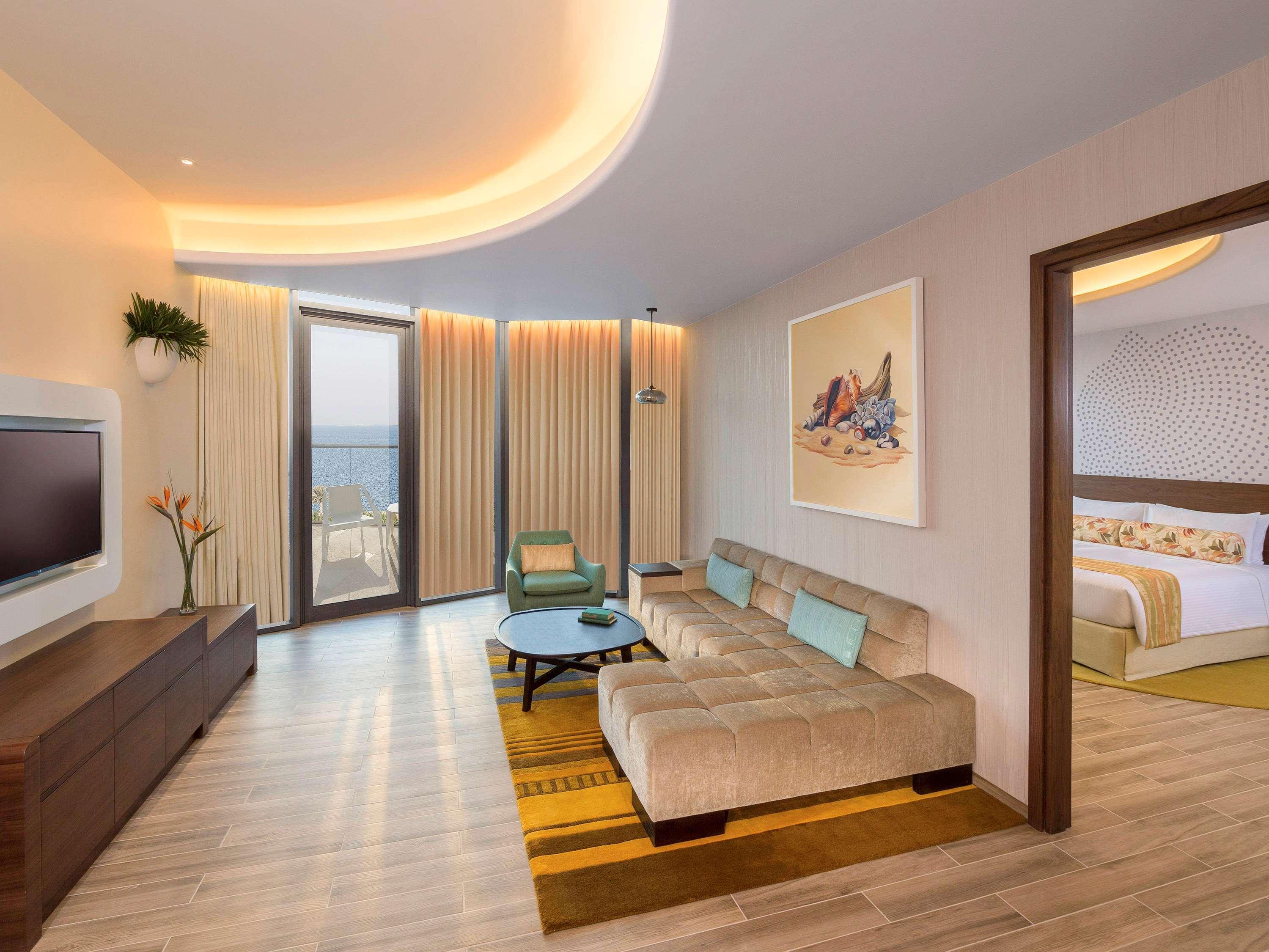 The Retreat Palm Dubai Mgallery By Sofitel Esterno foto The photo shows a modern hotel room or living area. It features a light-colored, spacious layout with a cozy, beige sofa placed in front of a low coffee table. There is a television mounted on the wall, and large windows with light-colored curtains t