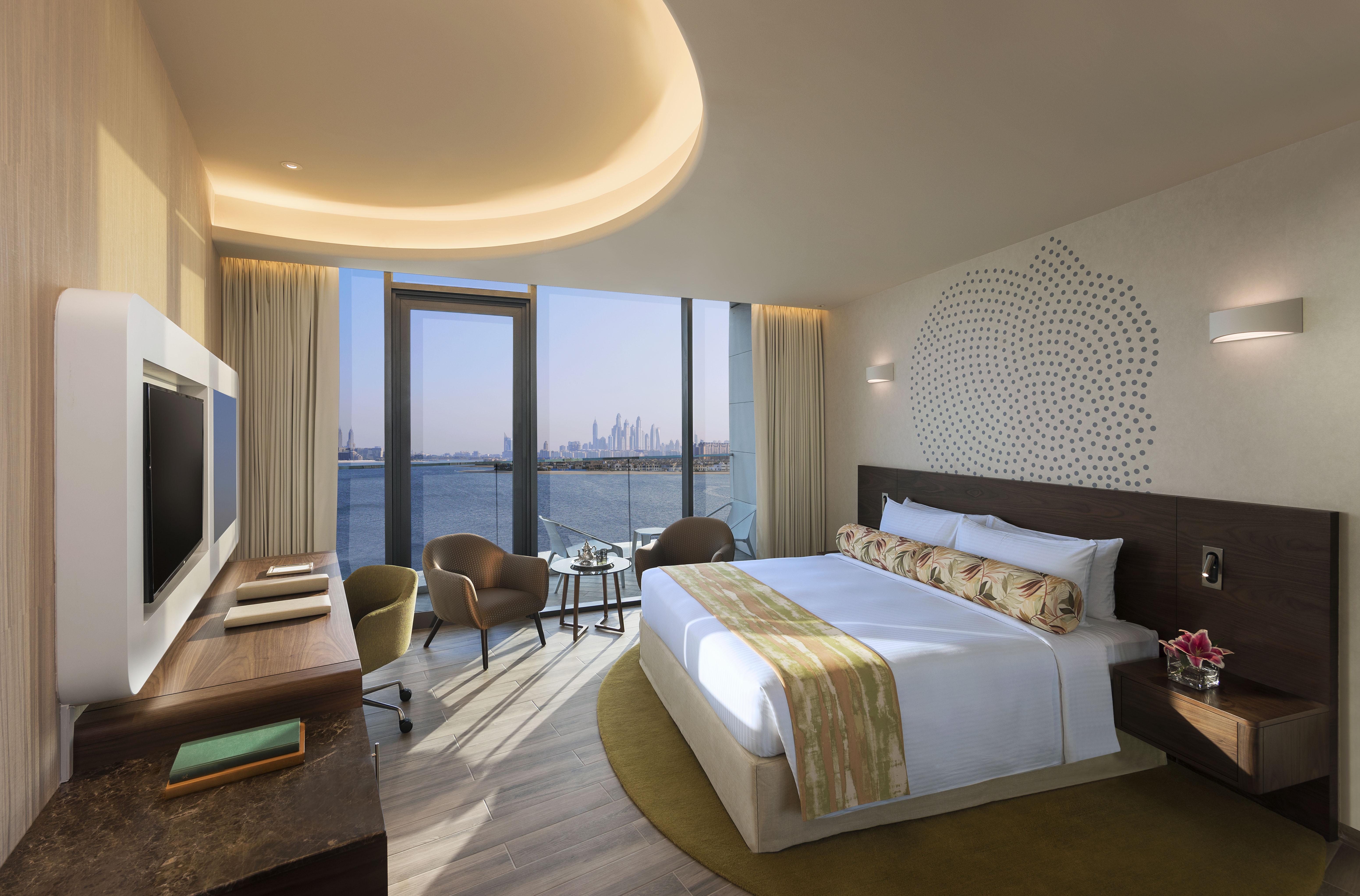 The Retreat Palm Dubai Mgallery By Sofitel Esterno foto The photo shows a modern hotel room featuring a large bed with decorative pillows and a plush headboard. The room is brightly lit with natural light coming through large windows that offer a scenic view of the water and the city skyline in the distan