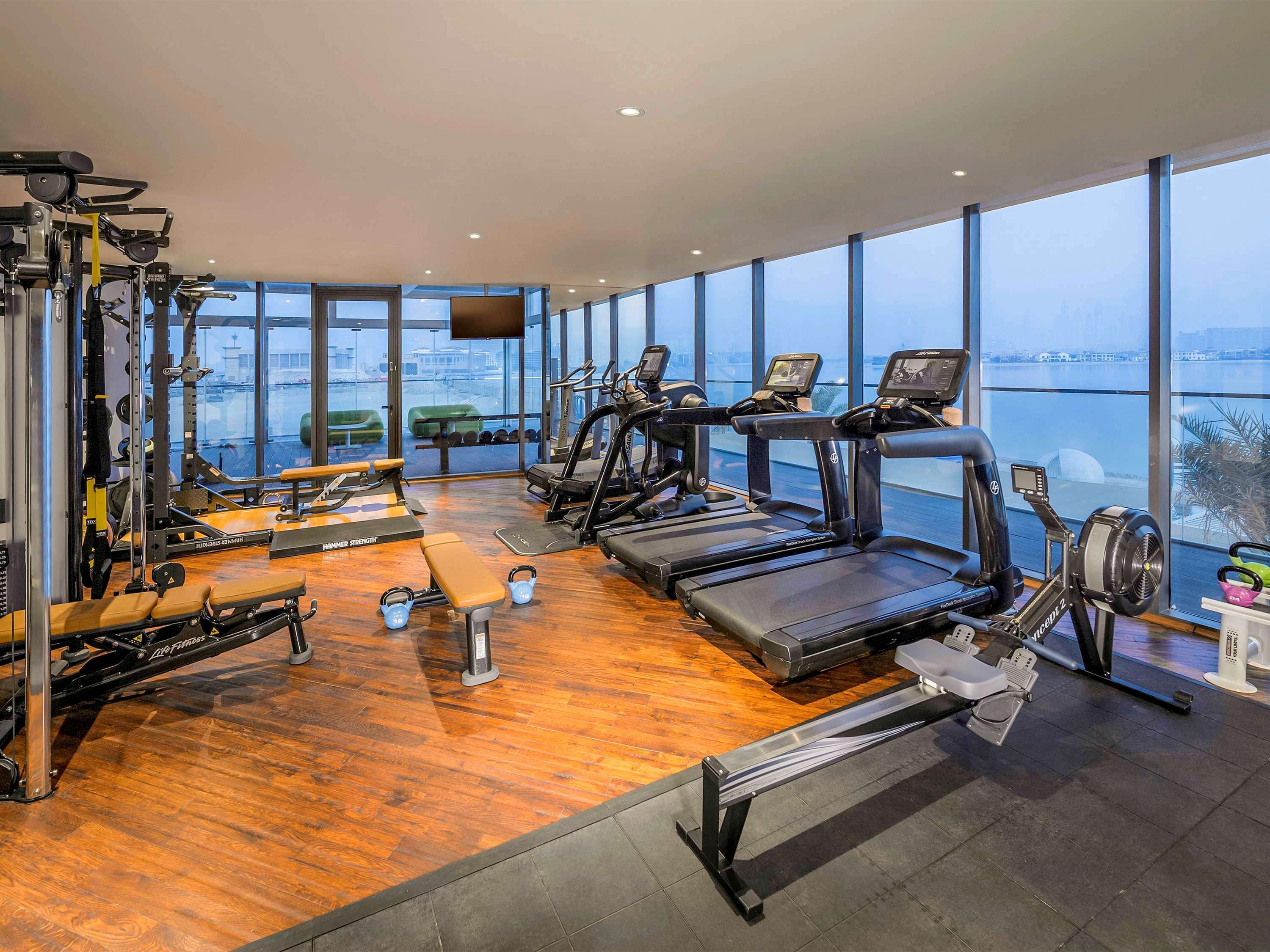 The Retreat Palm Dubai Mgallery By Sofitel Esterno foto The photo shows a modern gym with large windows that provide a view of the outside. The gym is equipped with various fitness equipment, including treadmills, rowing machines, and weight training equipment. The flooring is a combination of wood and ru