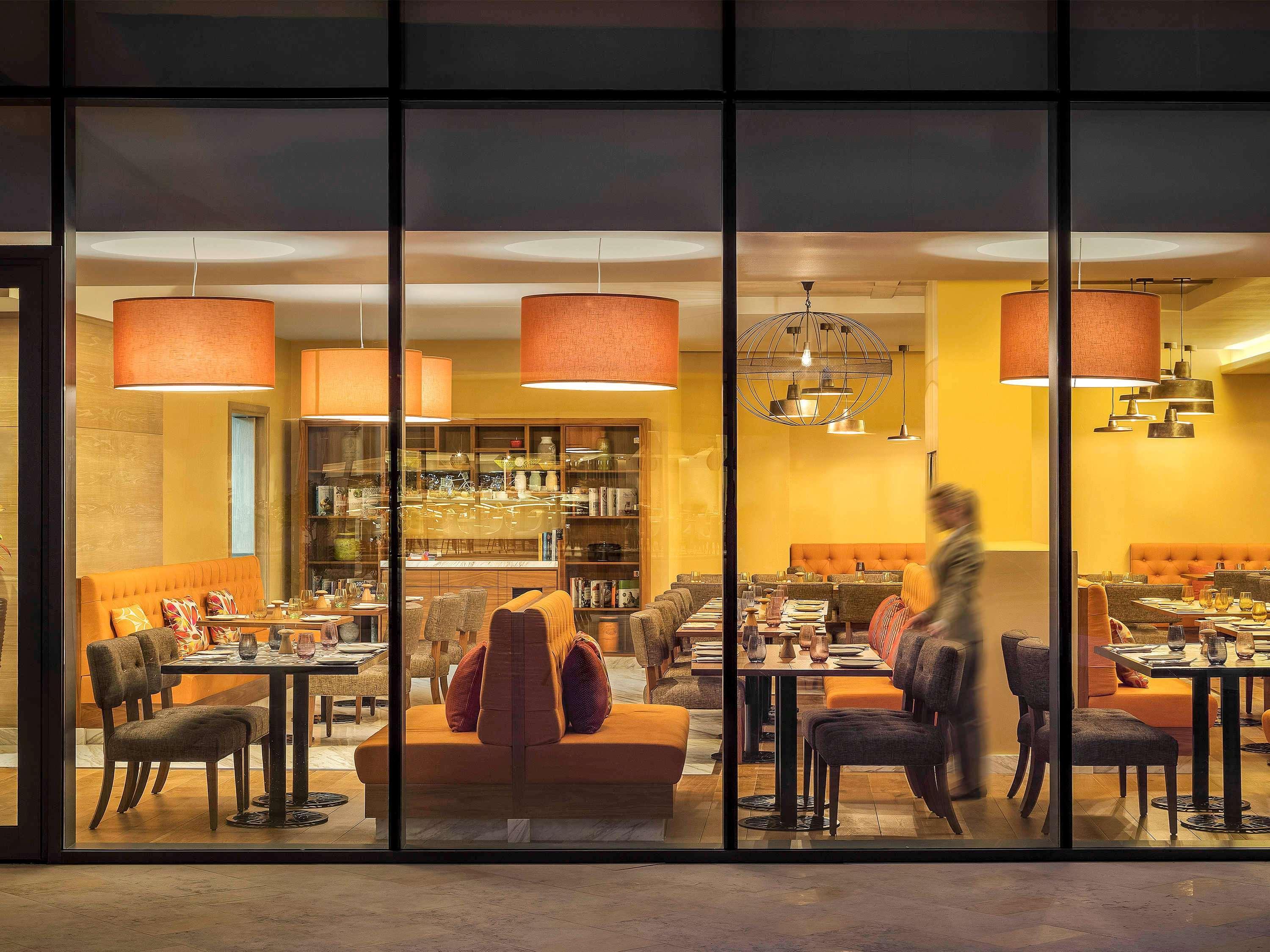 The Retreat Palm Dubai Mgallery By Sofitel Esterno foto The photo depicts a modern restaurant interior viewed through large glass windows. Inside, there are tables set for dining, with a mix of chairs and seating arrangements. The color scheme features warm, inviting tones, including yellow walls and soft