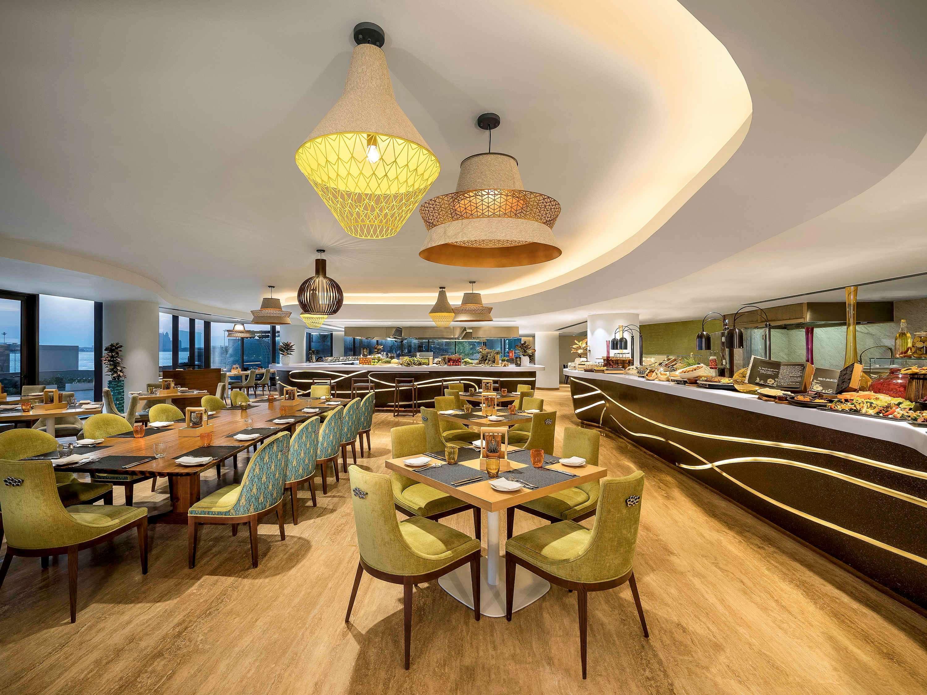 The Retreat Palm Dubai Mgallery By Sofitel Esterno foto The photo shows the interior of a modern restaurant. It features an elegant design with a curved ceiling that has stylish lighting fixtures, including hanging lamps in various shapes. The dining area consists of several round and rectangular wooden t