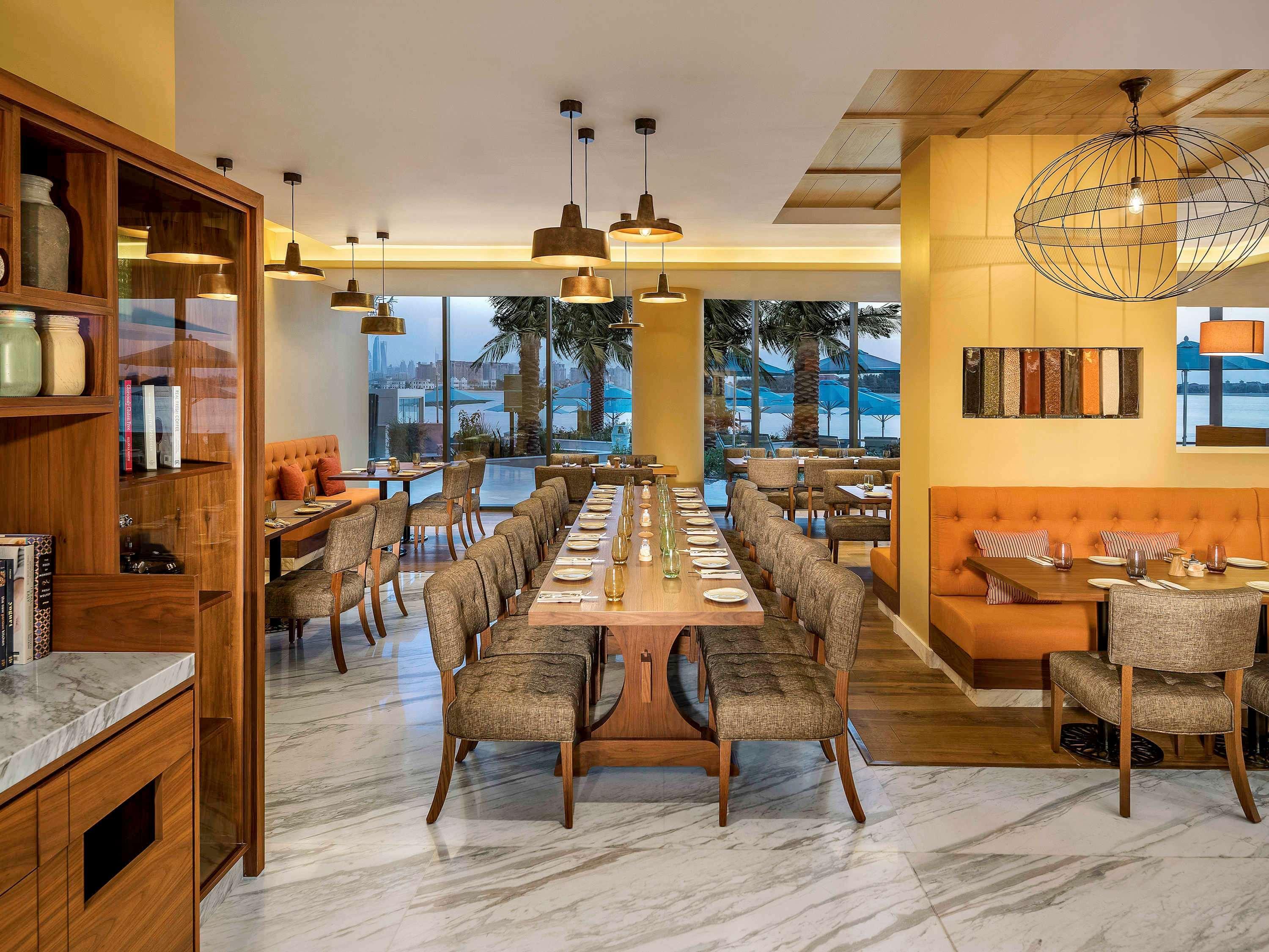 The Retreat Palm Dubai Mgallery By Sofitel Esterno foto The image shows a modern restaurant interior. It features a mix of wooden and marble elements, with a long dining table surrounded by upholstered chairs. The lighting is warm, with elegant pendant lights hanging from the ceiling. Large windows provid