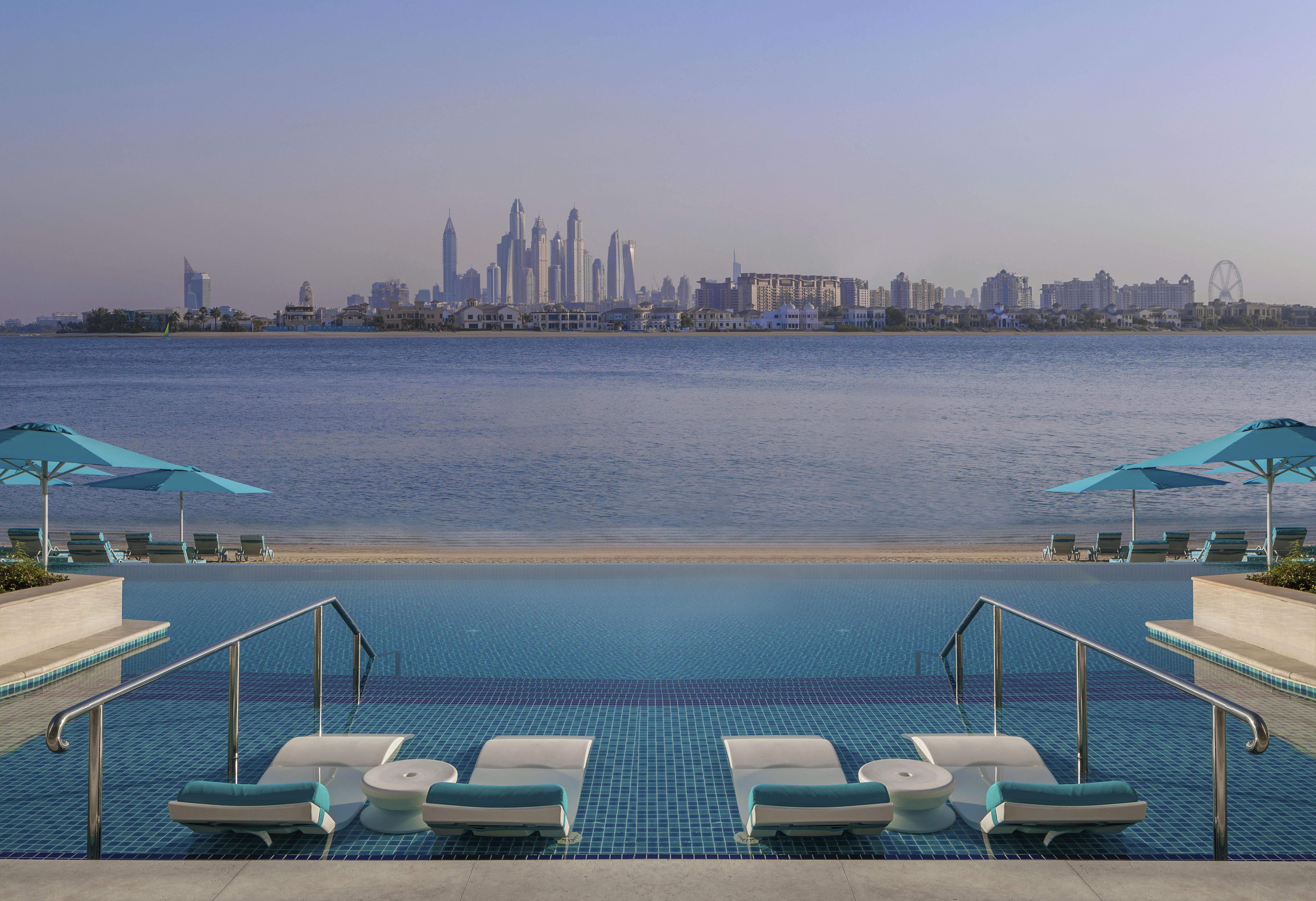 The Retreat Palm Dubai Mgallery By Sofitel Esterno foto The image shows a serene waterfront view featuring a luxurious pool area. In the foreground, there are several sun loungers and umbrellas, suggesting a relaxation space. The pool has a striking blue color, and its edge appears to blend into the body 