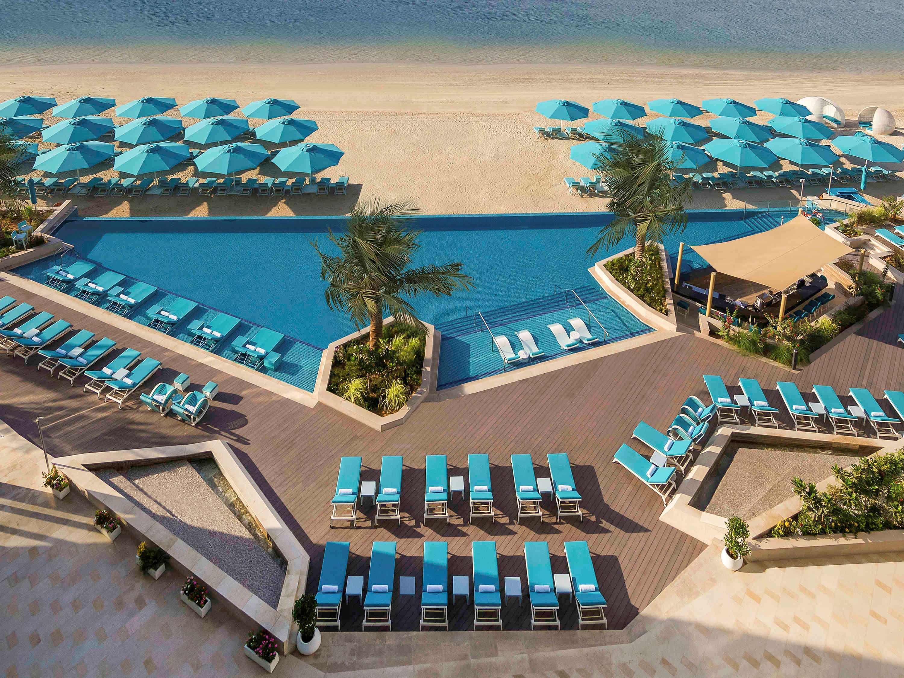 The Retreat Palm Dubai Mgallery By Sofitel Esterno foto The photo depicts a luxurious beachside resort area. In the foreground, there are several lounge chairs arranged neatly around a rectangular swimming pool. The pool is surrounded by lush greenery and palm trees, adding to the tropical ambiance. 

Fur