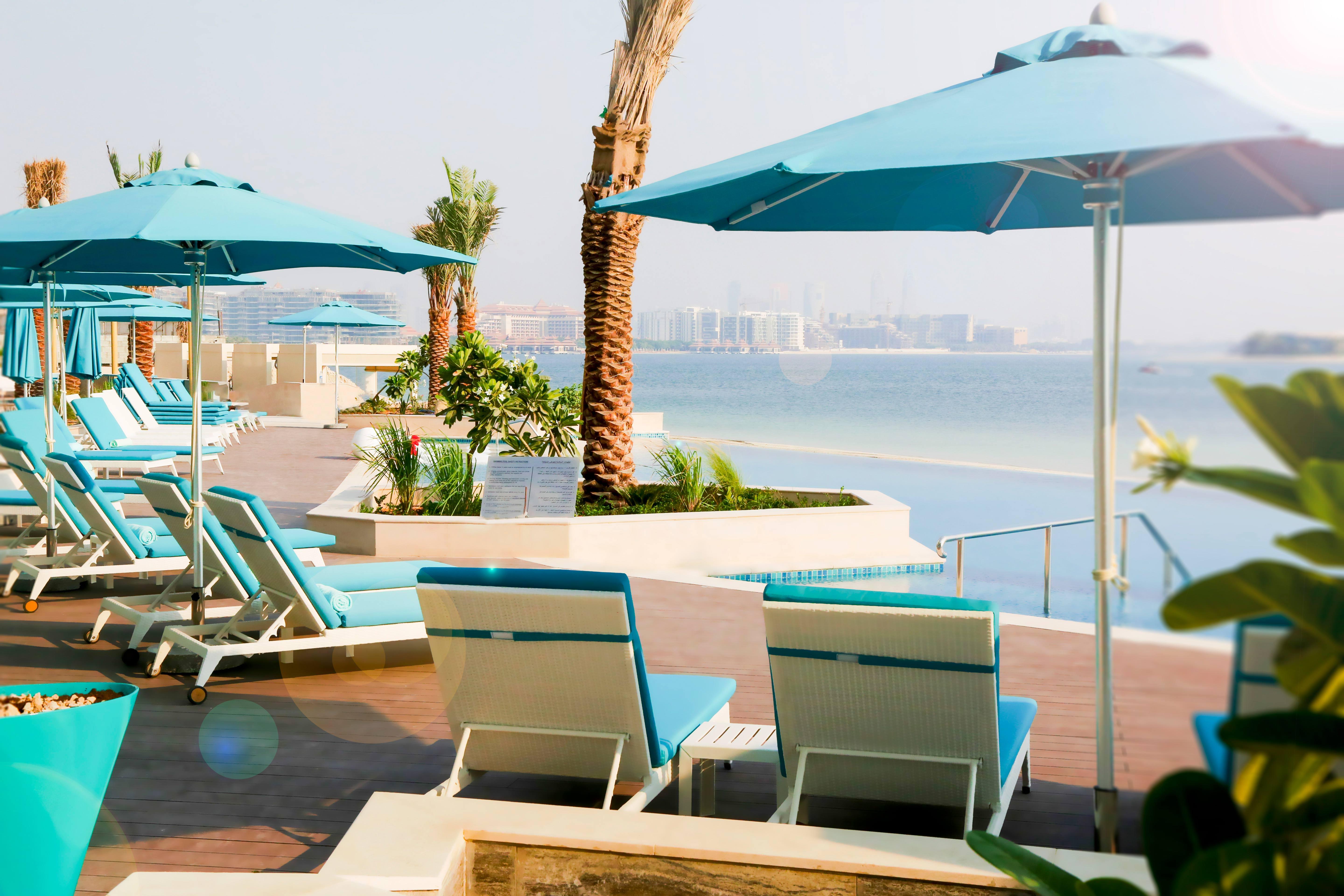 The Retreat Palm Dubai Mgallery By Sofitel Esterno foto The photo showcases a stylish outdoor lounge area at a resort. There are several light turquoise loungers arranged around a pool, each equipped with matching umbrellas. Palm trees and greenery add a tropical feel, while the calm sea can be seen in th