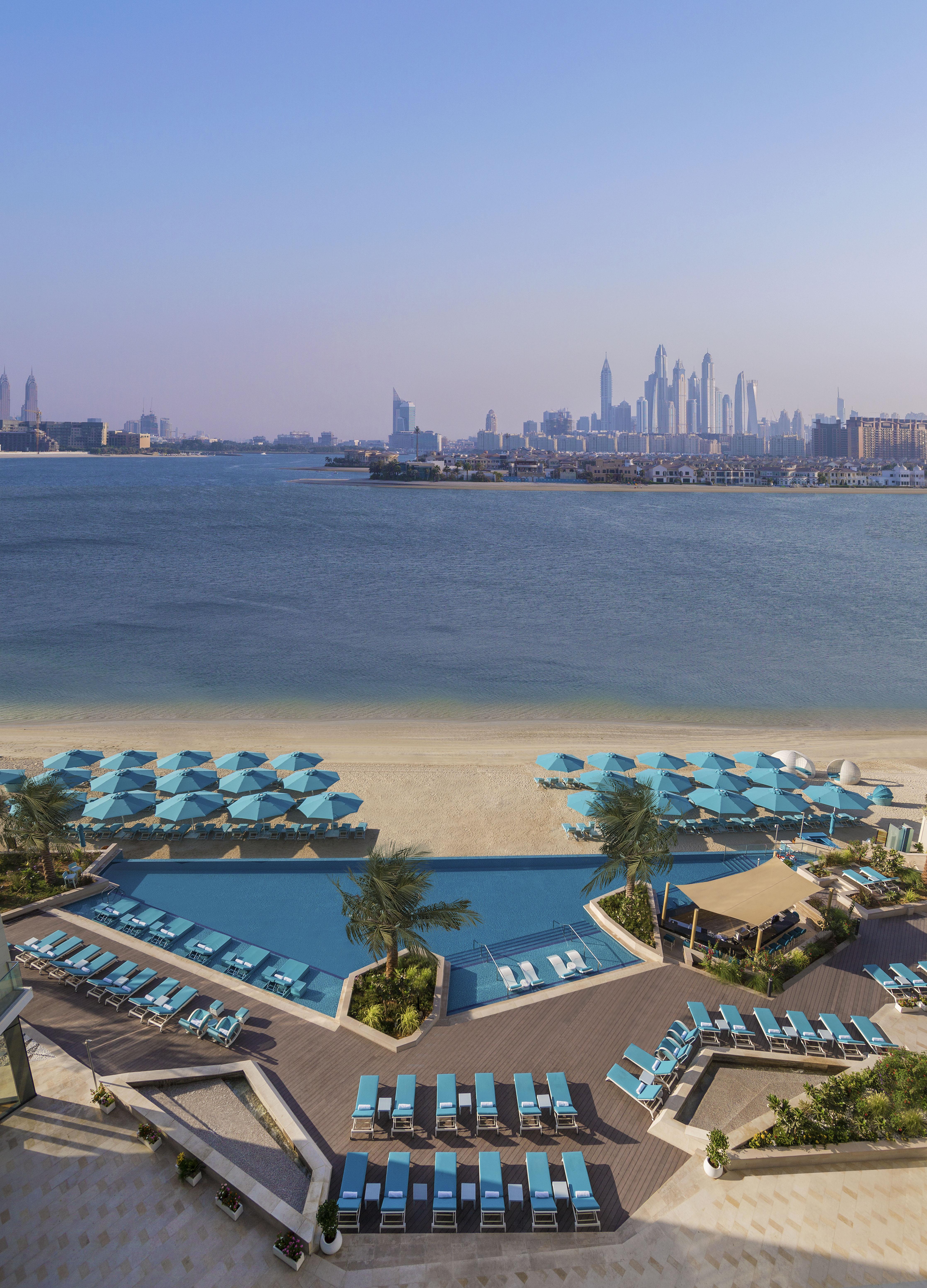 The Retreat Palm Dubai Mgallery By Sofitel Esterno foto The photo shows a scenic view of a beachfront resort area. In the foreground, there is a large swimming pool lined with sun loungers and umbrellas in shades of blue. Beyond the pool, there's a sandy beach leading to a body of water. In the background