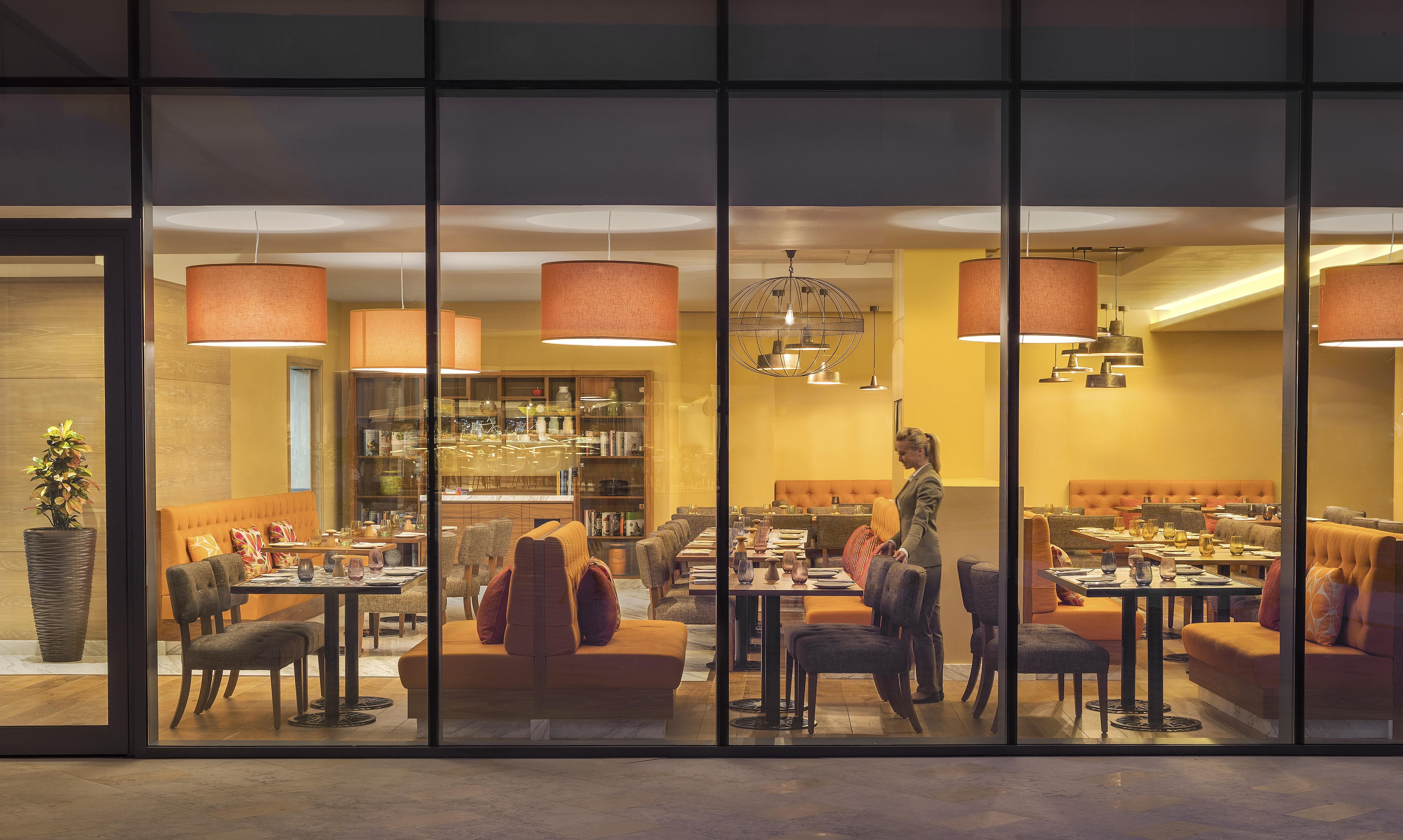 The Retreat Palm Dubai Mgallery By Sofitel Esterno foto The photo shows a modern restaurant interior as viewed through large glass windows. Inside, there are several tables and chairs arranged, with some featuring orange upholstered seating. The decor includes stylish pendant lights hanging from the ceili