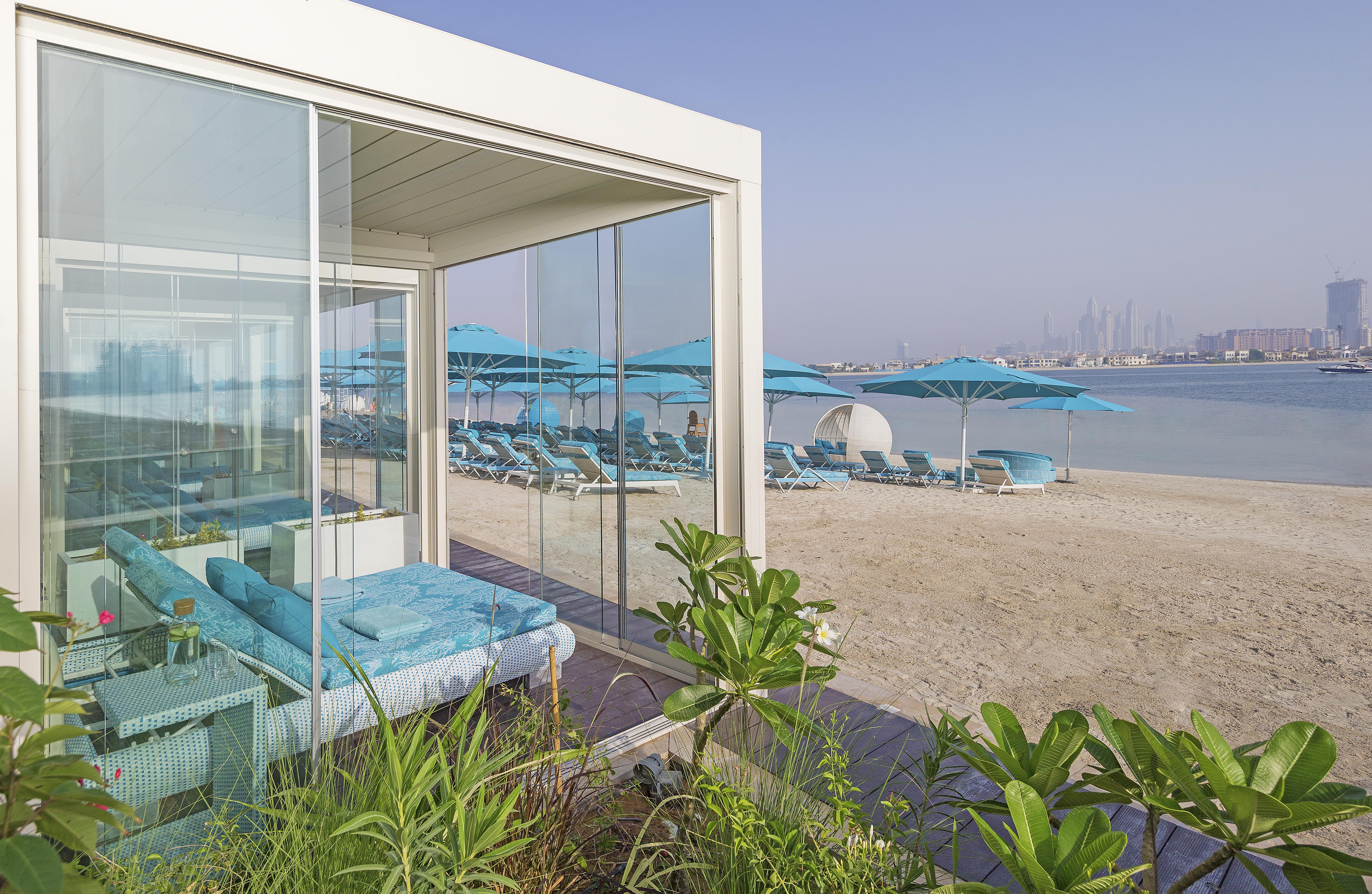 The Retreat Palm Dubai Mgallery By Sofitel Esterno foto The photo shows a modern, stylish beachfront setting. In the foreground, there is a glass-walled structure that appears to be a lounge or cabana, featuring comfortable blue seating. Beyond the structure, there is a sandy beach with several turquoise 