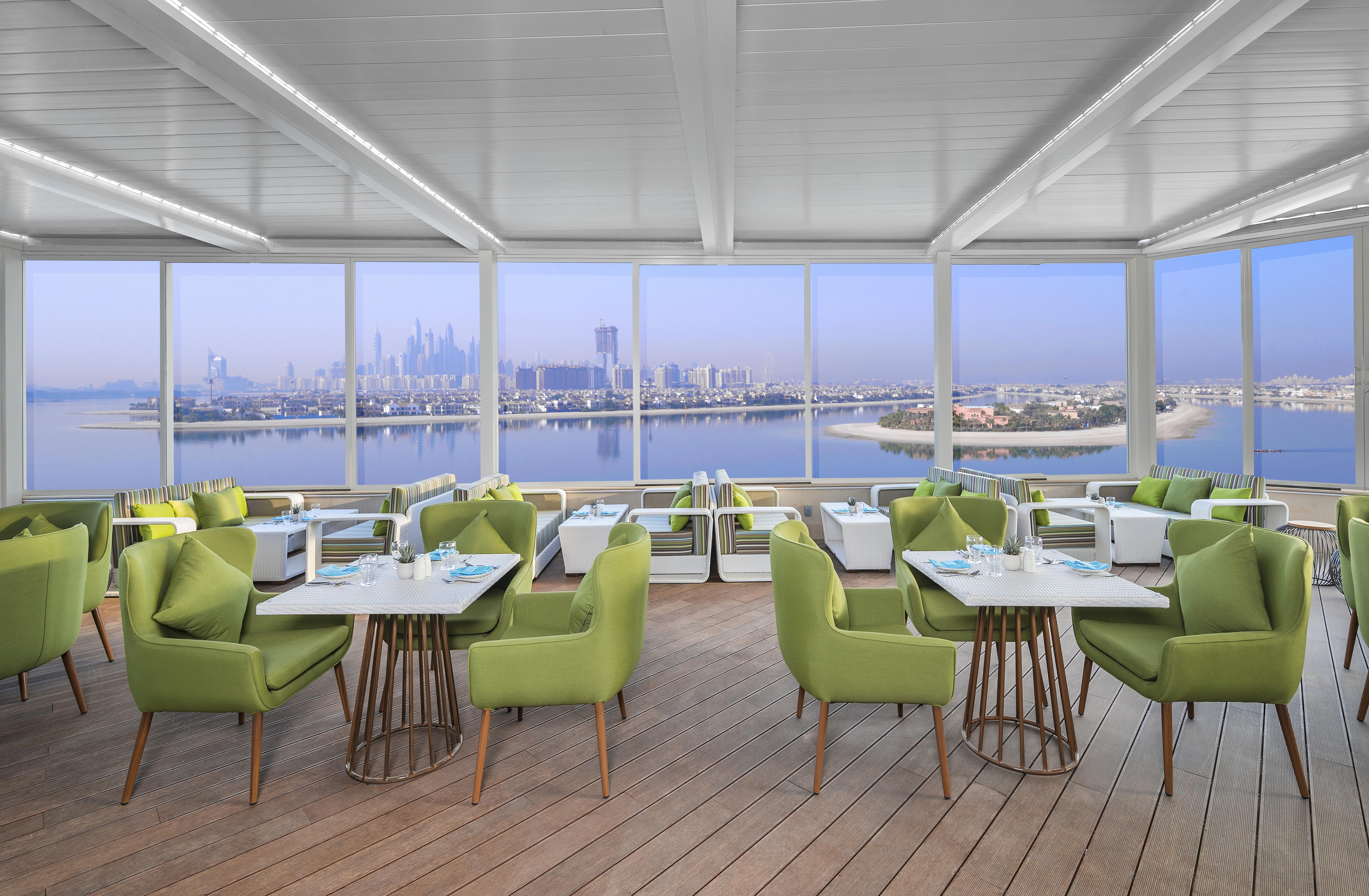 The Retreat Palm Dubai Mgallery By Sofitel Esterno foto The photo shows an elegant, modern restaurant or café with large windows that provide a panoramic view of a city skyline and waterfront. The interior features green chairs with wooden legs arranged around white tables. The floor is made of light wood