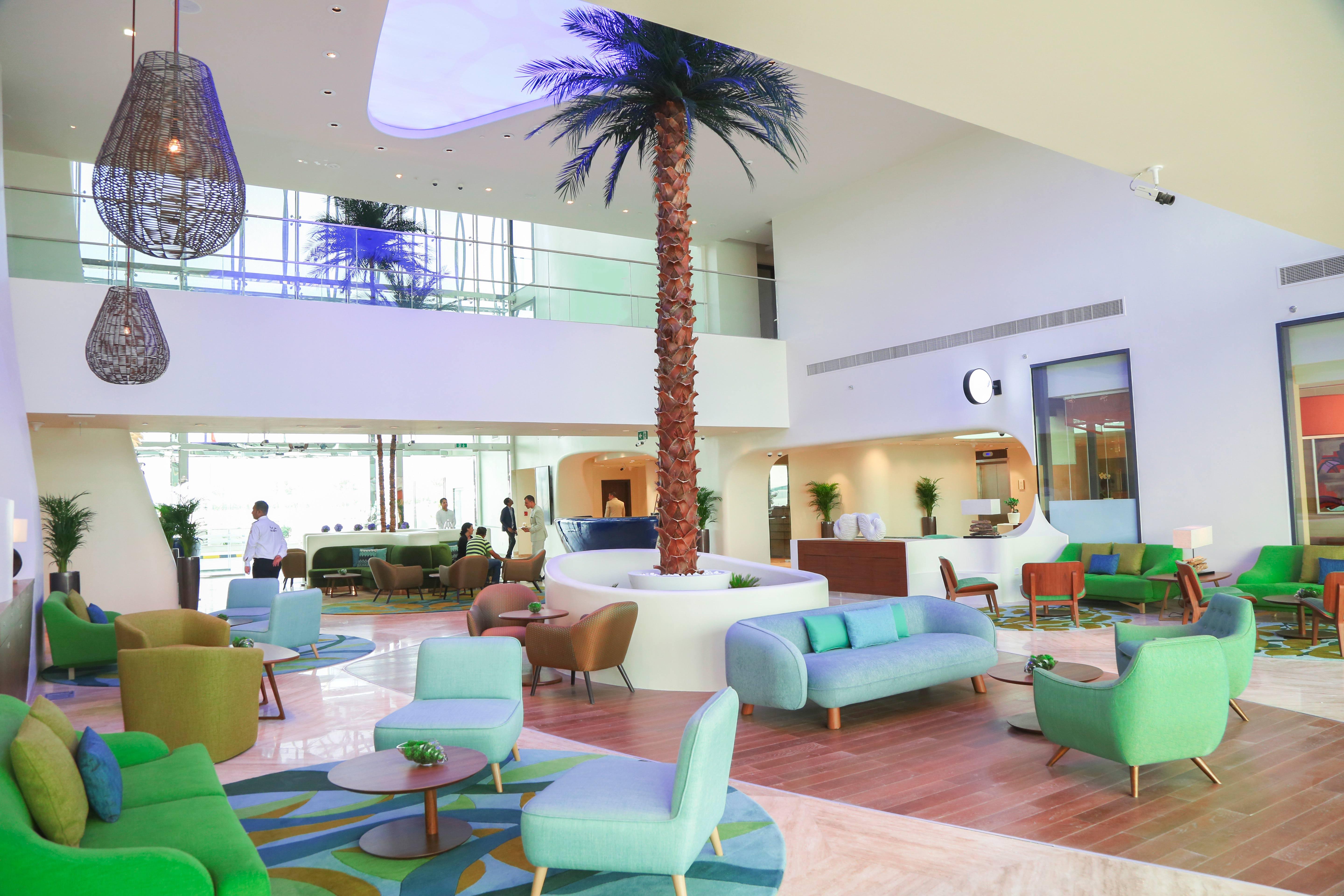 The Retreat Palm Dubai Mgallery By Sofitel Esterno foto The photo shows a modern and stylish lobby area. It features a high ceiling with large windows allowing natural light to fill the space. In the center, there is a tall palm tree, adding a tropical feel. The seating consists of colorful couches and ch