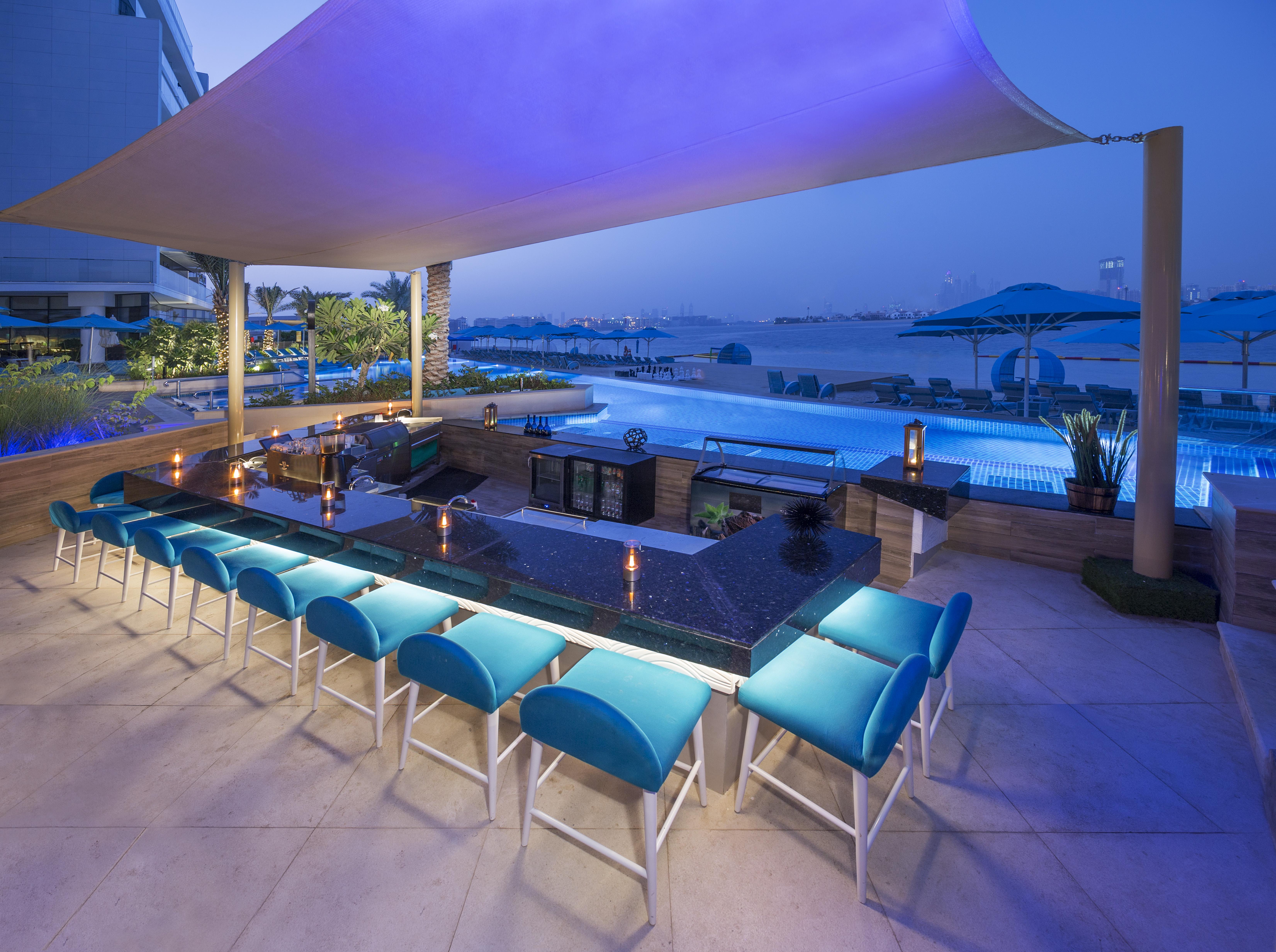 The Retreat Palm Dubai Mgallery By Sofitel Esterno foto The photo depicts an elegant outdoor bar area beside a swimming pool. It features a long, dark countertop with several bar stools that have vibrant blue cushions. The setting is illuminated with soft lighting, creating a relaxed and inviting atmosphe