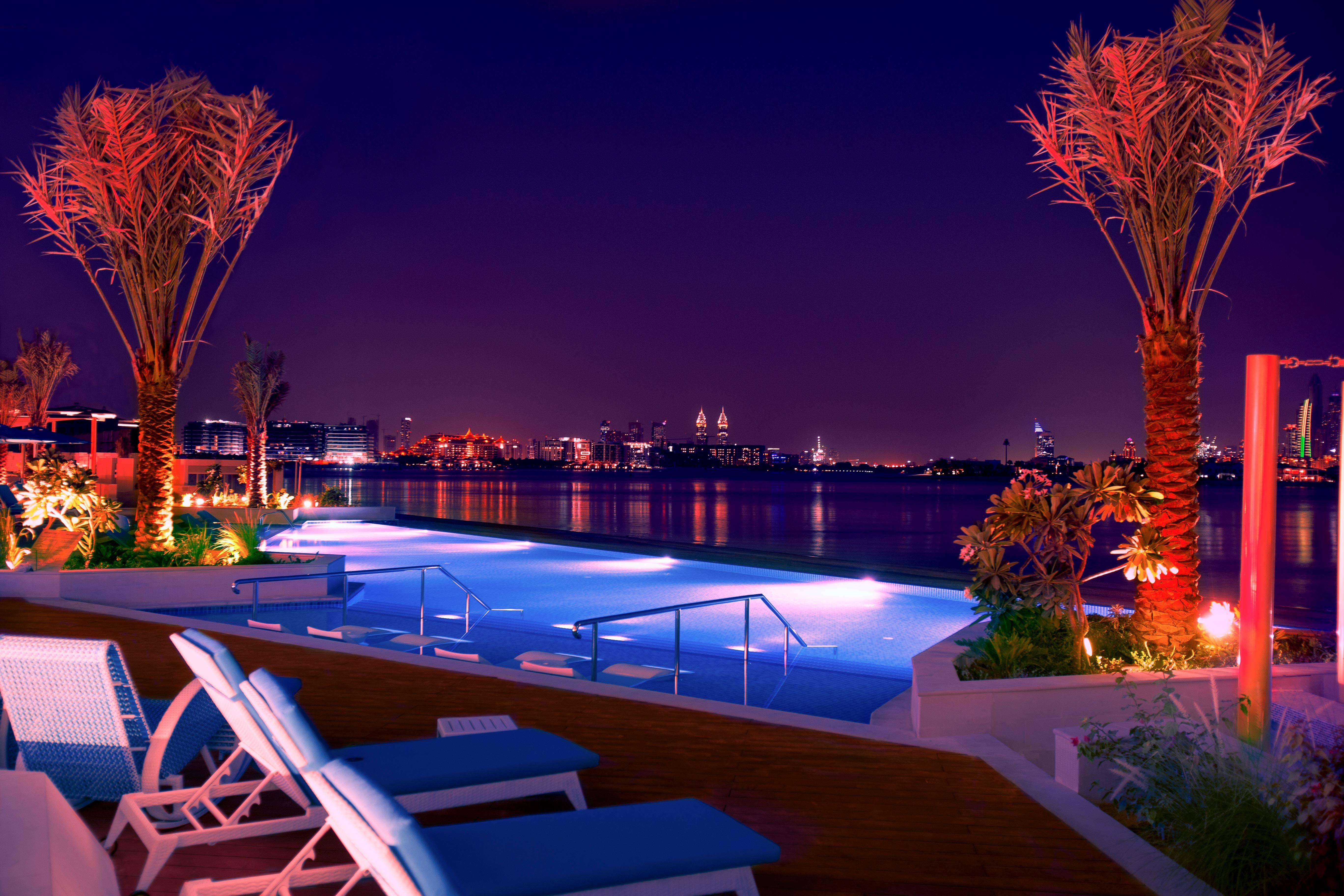 The Retreat Palm Dubai Mgallery By Sofitel Esterno foto The photo depicts a luxurious outdoor pool area at night. The pool is illuminated with soft blue and pink lights, creating a serene atmosphere. Surrounding the pool are lounge chairs, inviting relaxation. Palm trees with vibrant lighting add to the t