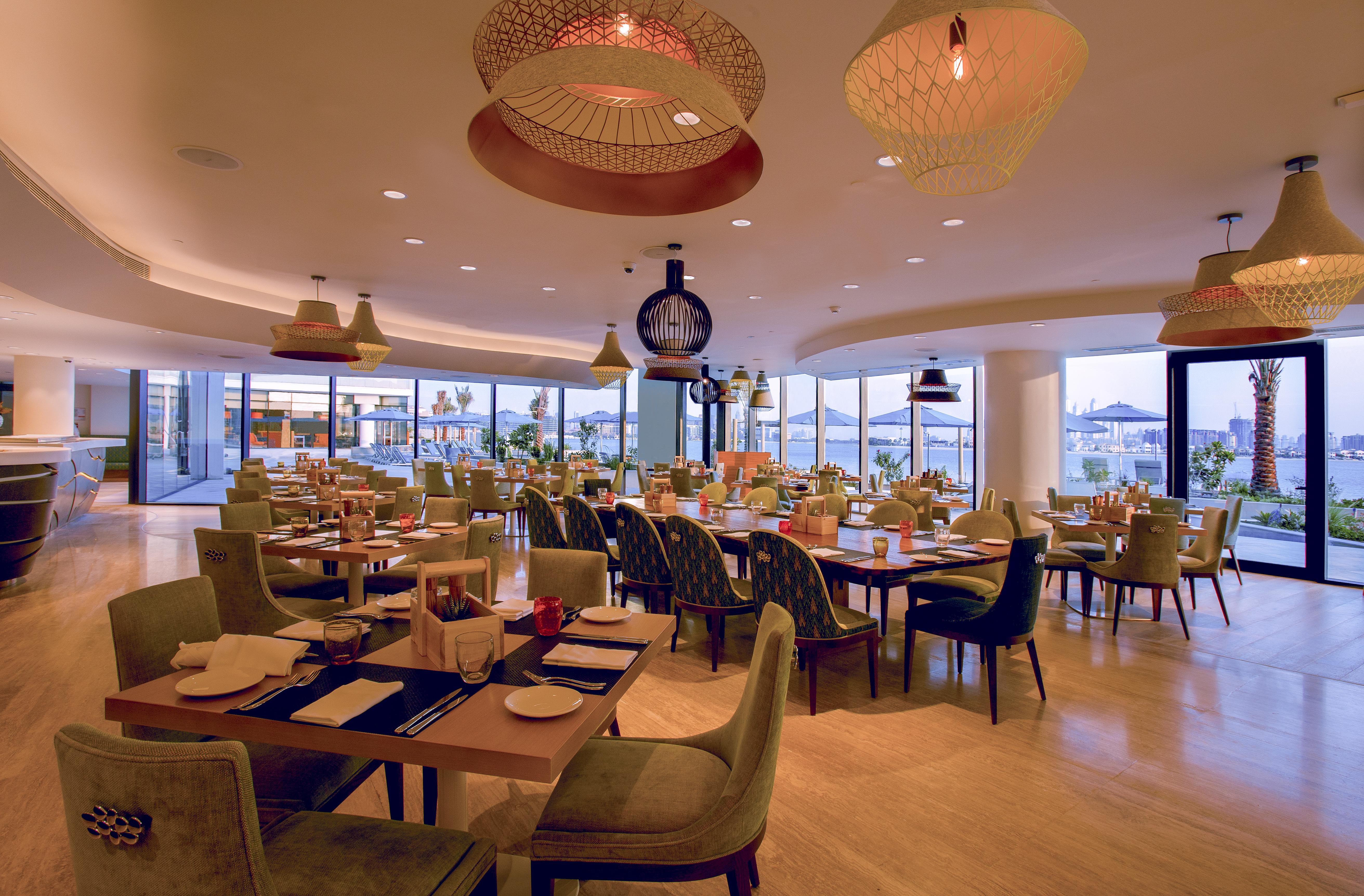 The Retreat Palm Dubai Mgallery By Sofitel Esterno foto The photo shows an elegant restaurant interior. It features several tables arranged for dining, with stylish chairs and neatly set tables. The decor includes modern lighting fixtures, including pendant lights with a woven design. Large windows provid