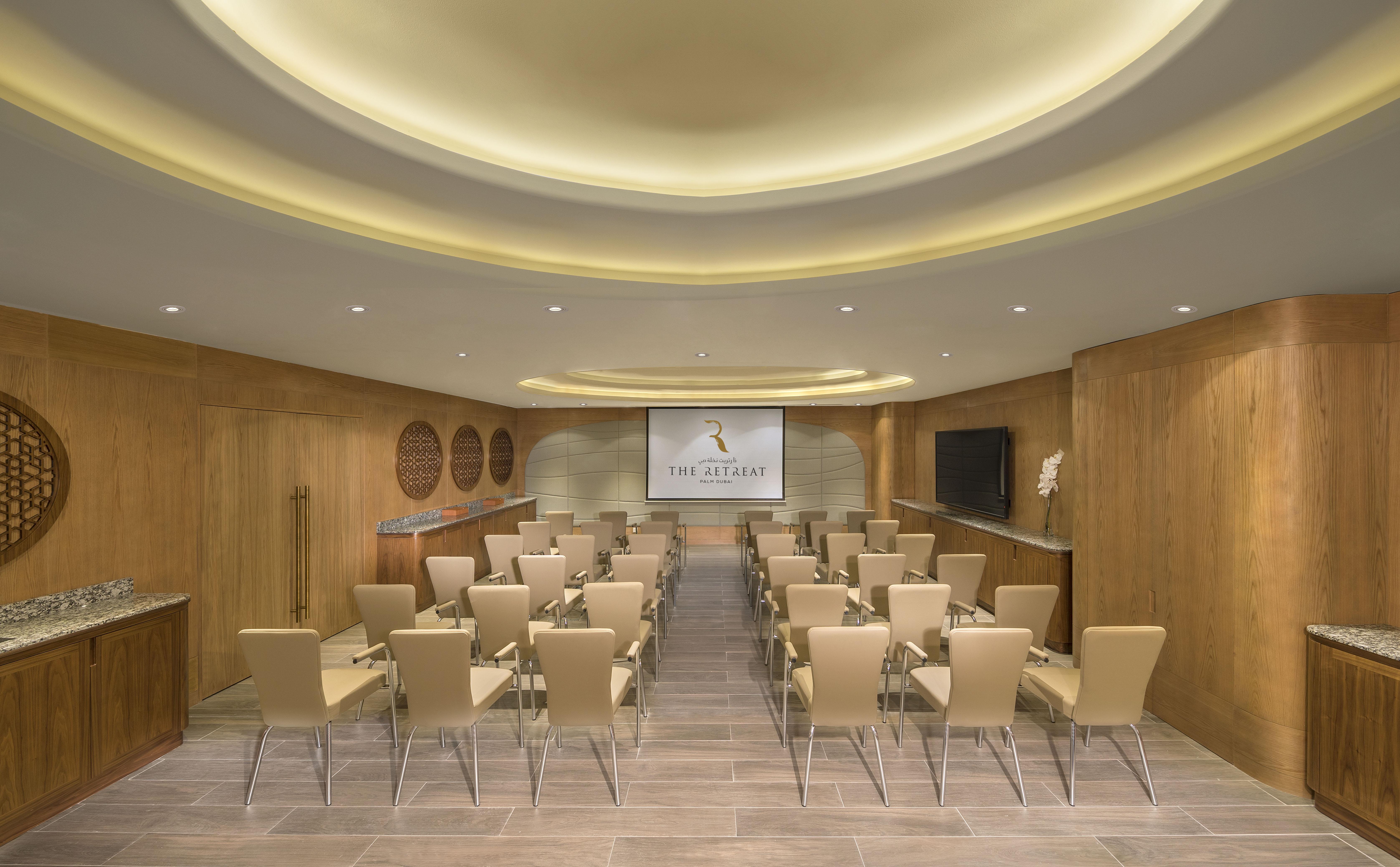 The Retreat Palm Dubai Mgallery By Sofitel Esterno foto The photo shows a modern conference room. The room features a light wood and neutral decor with a curved ceiling that has soft lighting. There are rows of beige chairs arranged neatly facing a large screen on one wall. A sleek, wooden side area appea