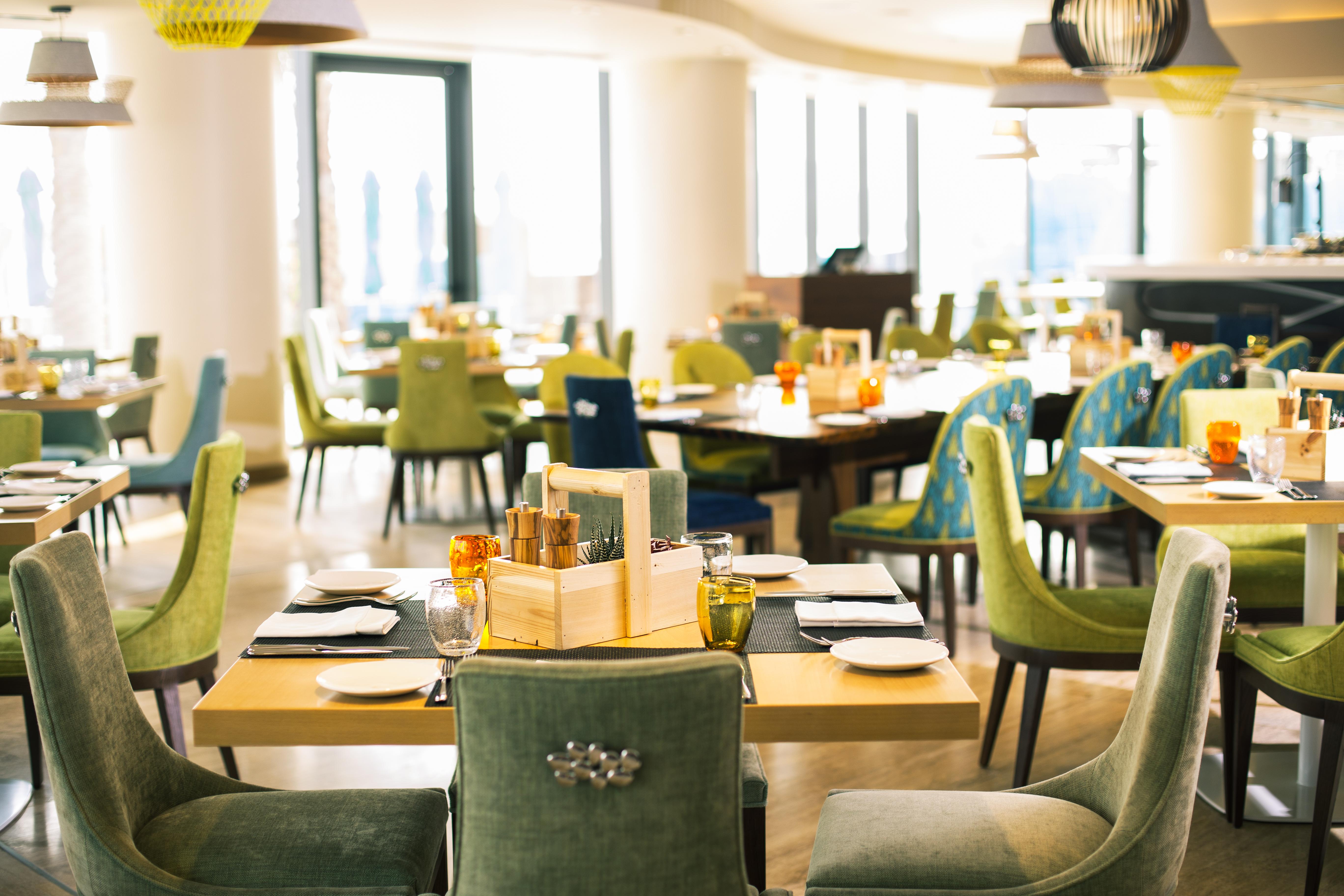 The Retreat Palm Dubai Mgallery By Sofitel Esterno foto The photo shows a bright and modern restaurant interior. There are several dining tables set with cutlery and plates. The chairs come in various colors, primarily green and blue, creating a vibrant atmosphere. Large windows allow natural light to fil
