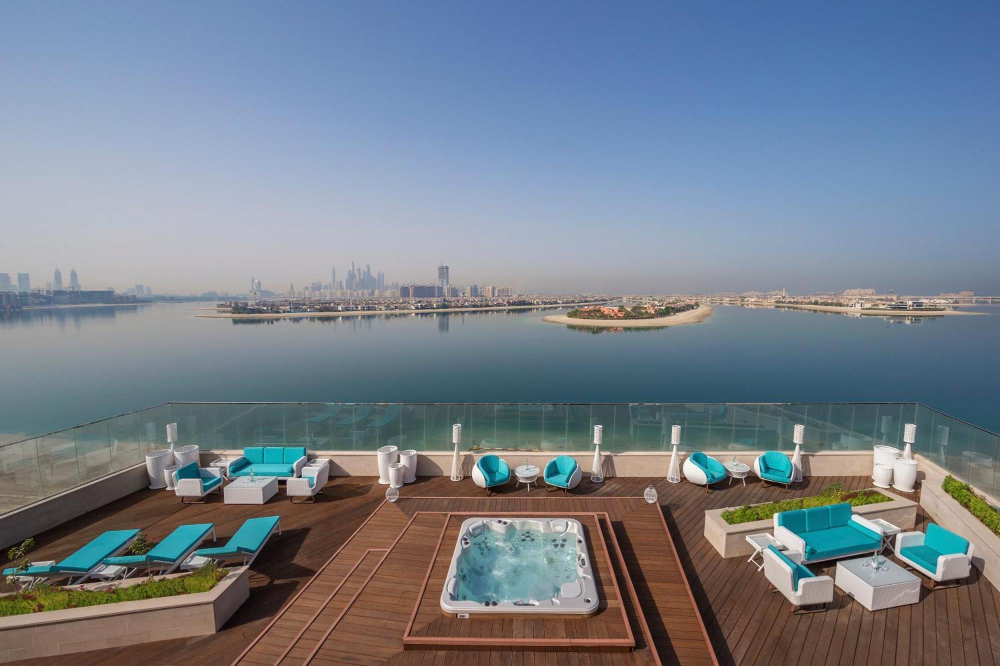 The Retreat Palm Dubai Mgallery By Sofitel Esterno foto The photo showcases a luxurious outdoor area, likely a rooftop terrace or poolside setting. There are several sun loungers arranged neatly, featuring turquoise cushions and a wooden deck. In the center, there is a hot tub or Jacuzzi, inviting for rel