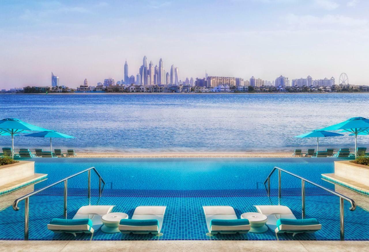 The Retreat Palm Dubai Mgallery By Sofitel Esterno foto The photo shows a scenic view of a luxurious pool area leading into a serene body of water. In the foreground, there are several white lounge chairs with turquoise cushions, and the pool features a modern design with steps leading into the water. The