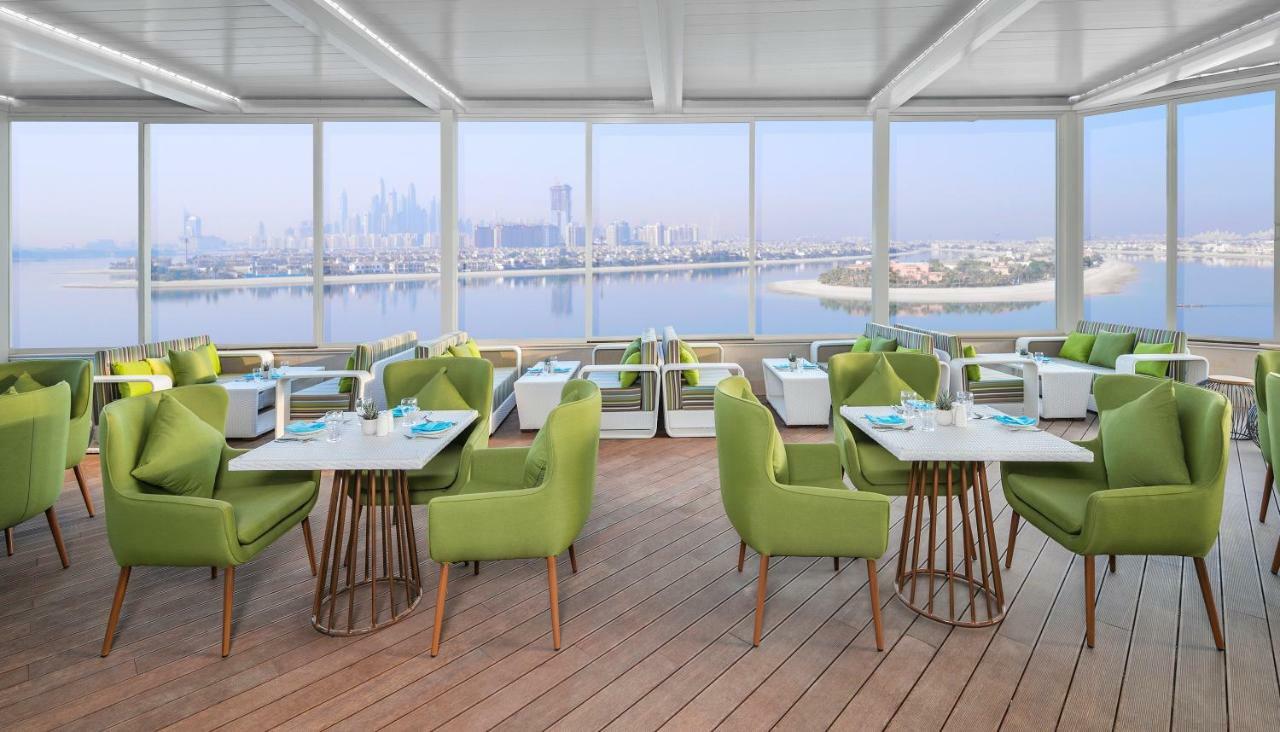 The Retreat Palm Dubai Mgallery By Sofitel Esterno foto The photo shows a modern dining area with a scenic view. It features several tables set with light-colored tableware and napkins. The chairs are upholstered in a vibrant green fabric, and the flooring appears to be wooden, contributing to a warm and 