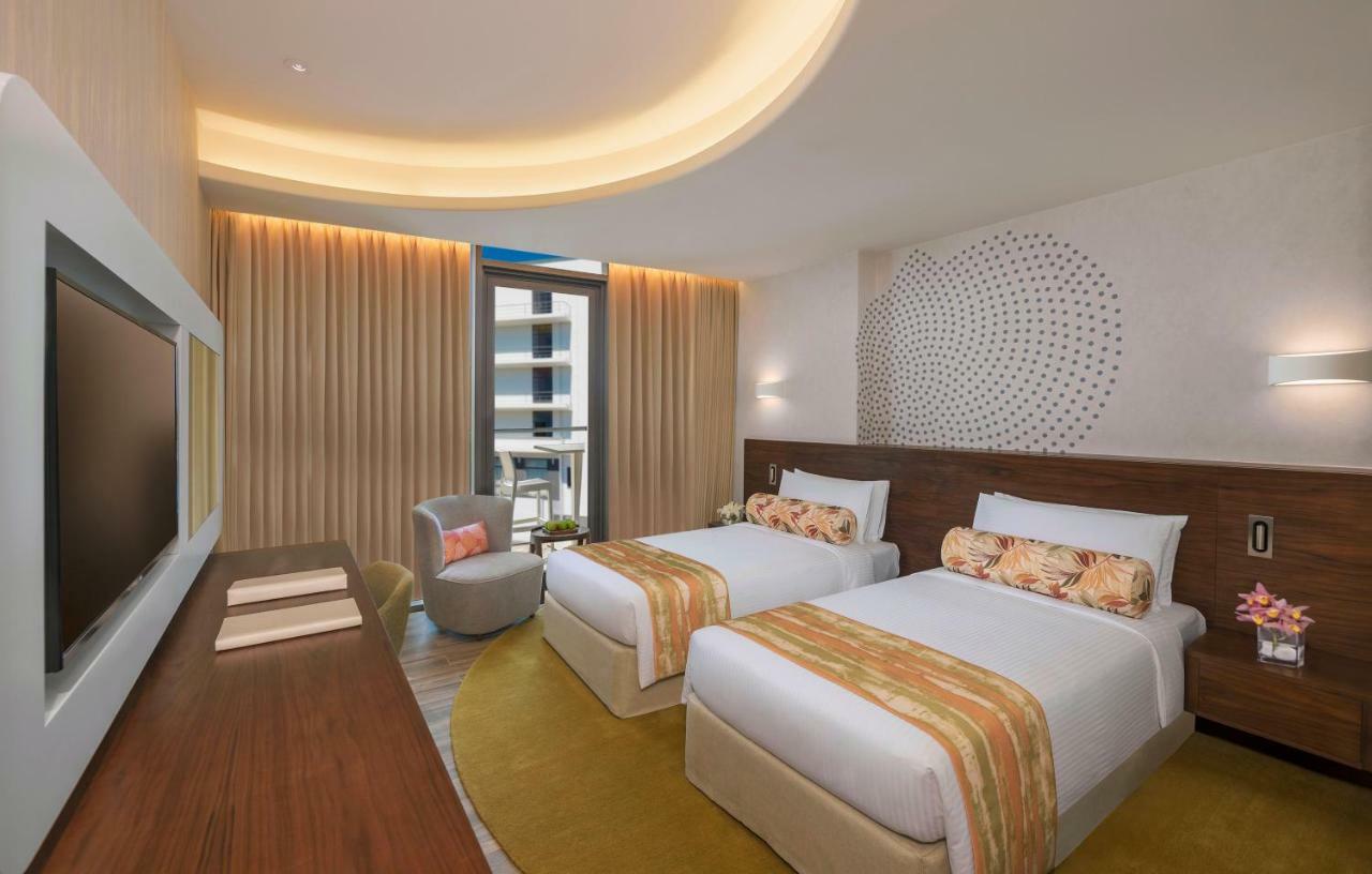 The Retreat Palm Dubai Mgallery By Sofitel Esterno foto The photo shows a hotel room featuring two single beds adorned with decorative cushions. The room has a modern design, with soft lighting and warm tones. There is a comfortable chair in the corner and a large television mounted on the wall. Floor-to-