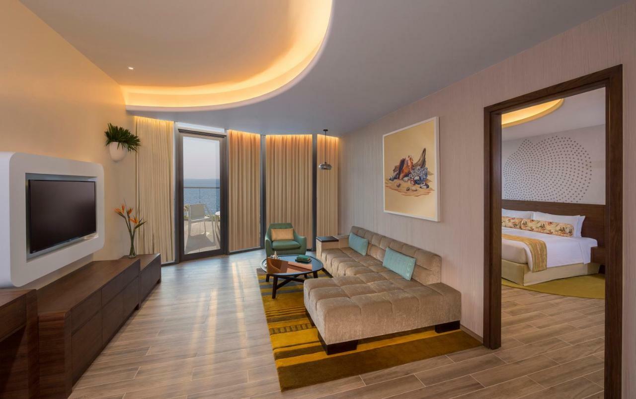 The Retreat Palm Dubai Mgallery By Sofitel Esterno foto The photo shows a modern hotel room or living space. It features a spacious layout with light wooden flooring and warm lighting. In the center, there's a comfortable, beige sofa with blue cushions. A small coffee table sits in front of it. To the lef